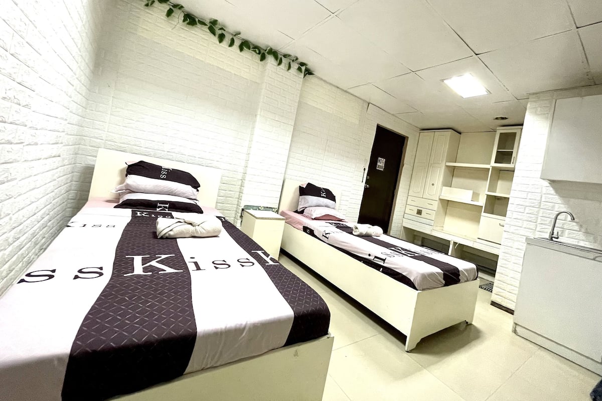 Ein Zimmer mit zwei Betten, jeweils mit einer Bettdecke von 'Kiss', einer weißen Ziegelwand, einem kleinen Fenster und einer Küchenzeile mit Spüle und Schränken.
