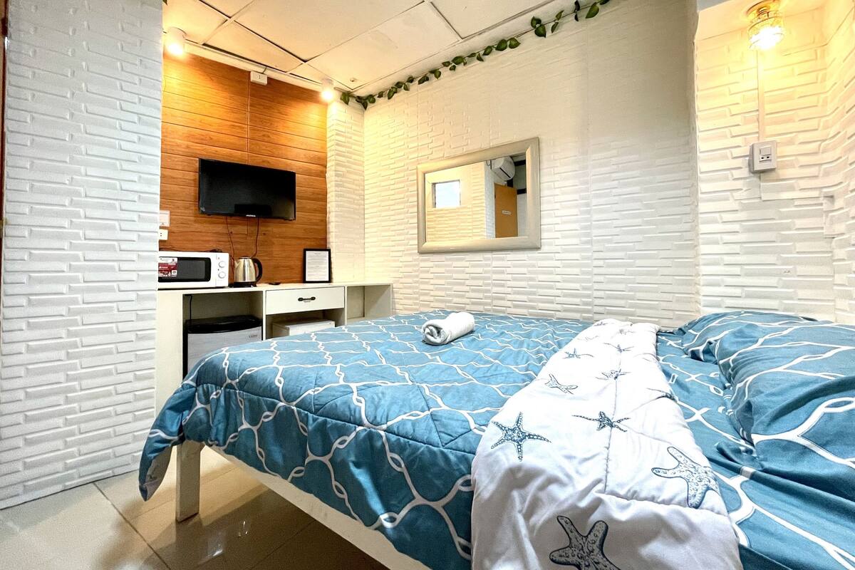 Ein Hotelzimmer mit einem Bett, einem an der Wand befestigten Fernseher, einem kleinen Schreibtisch mit Wasserkocher und einem Spiegel.