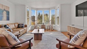 A modern living room with a leather sofa, wooden chairs, a coffee table, and a large window with blinds.