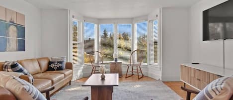 A modern living room with a leather sofa, wooden chairs, a coffee table, and a large window with blinds.