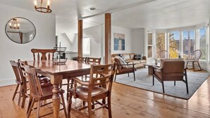 A spacious room with a wooden dining table, chairs, a sofa, and a coffee table. There are large windows with blinds, a mirror on the wall, and a chandelier.