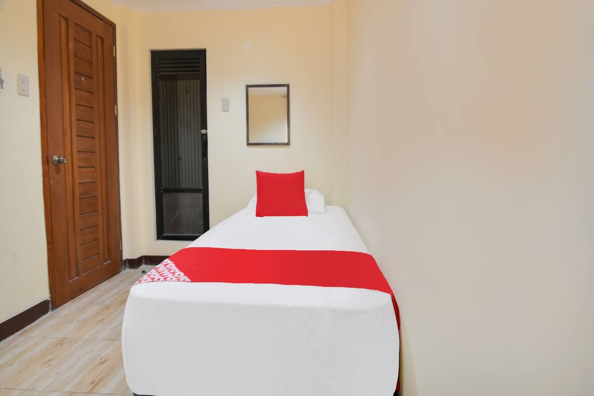 Un lit simple avec un oreiller rouge et du linge de lit blanc, dans une pièce aux murs de couleur beige et dotée d’une porte en bois.