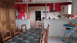 A dining area with a table set for six, wooden chairs, a baby high chair, a kitchen with white cabinets, a red wall, and a chandelier. - Spacious house with enclosed garden in Sainte-Mère-Église, pets allowed (Sainte-Mère-Église)