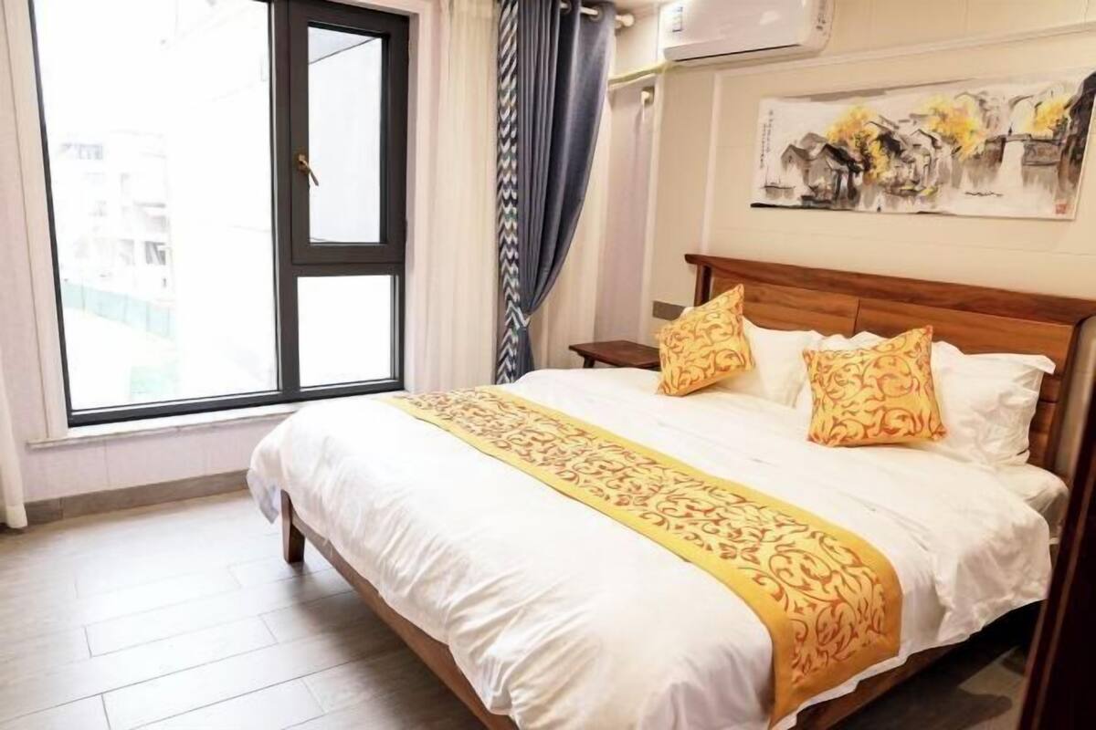 A hotel room with a large bed, a window with curtains, a painting on the wall, and a wooden nightstand.