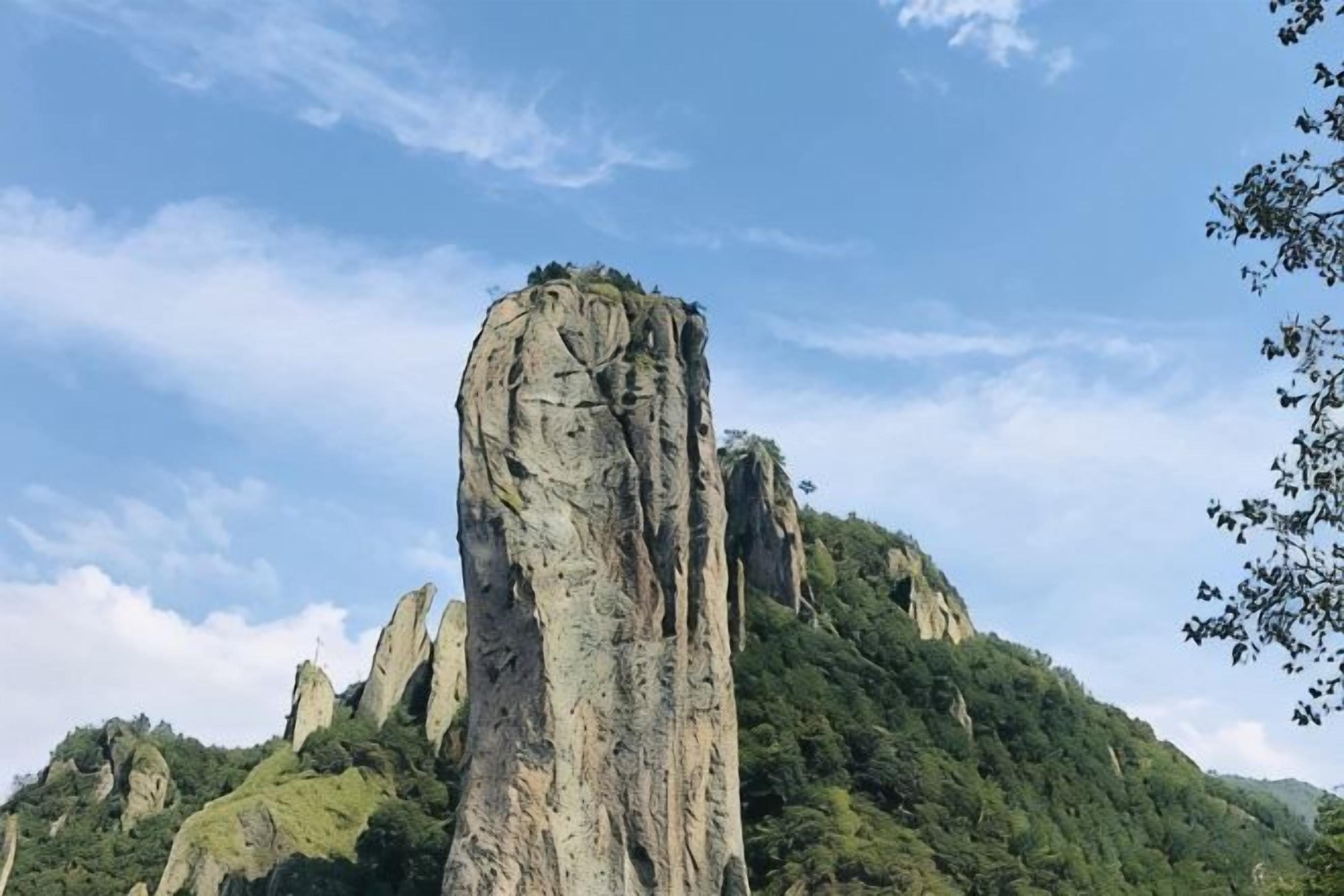 A tall, narrow rock formation with a forested hill in the background.