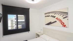 Een hotelkamer met een bed, een raam met uitzicht op gebouwen, een wandklok en een plafondventilator.