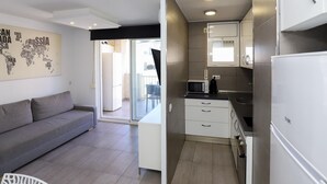 Een compacte keuken met witte kastjes, een magnetron en een koelkast. In de woonkamer staat een grijze bank en op de muur een wereldkaart.