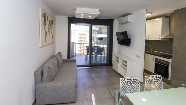 Een modern appartement met een grijze bank, een glazen eettafel en een keuken met witte kasten en een inbouwoven.