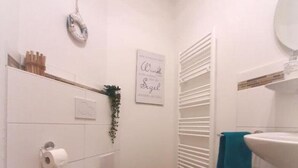 Une petite salle de bain blanche, avec des toilettes, un lavabo et un miroir. Une serviette est accrochée au lavabo, et il y a un panneau sur le mur.