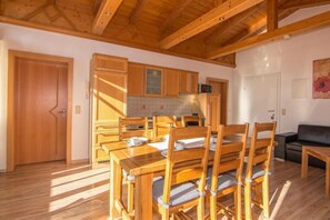 Una mesa de comedor de madera con sillas, una zona de cocina con armarios de madera y un sofá negro en una habitación con suelo de madera y techo inclinado.