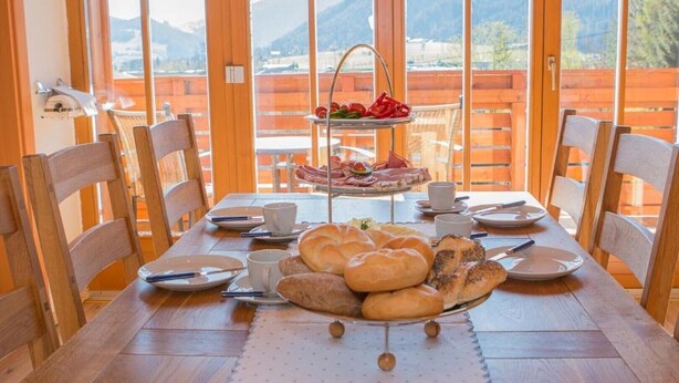 Una mesa de comedor servida con pan, pasteles y una pequeña bandeja de frutas, con vistas panorámicas a las montañas a través de amplios ventanales.