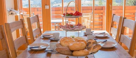 Una mesa de comedor servida con pan, pasteles y una pequeña bandeja de frutas, con vistas panorámicas a las montañas a través de amplios ventanales.