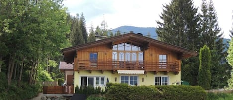 Un chalet de madera de dos pisos con un balcón, rodeado de vegetación exuberante y una montaña al fondo.