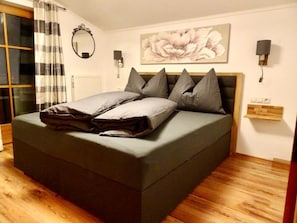 Een moderne slaapkamer met een groot bed, een houten vloer, een raam met gordijnen, een wandspiegel en een bloemrijk schilderij.