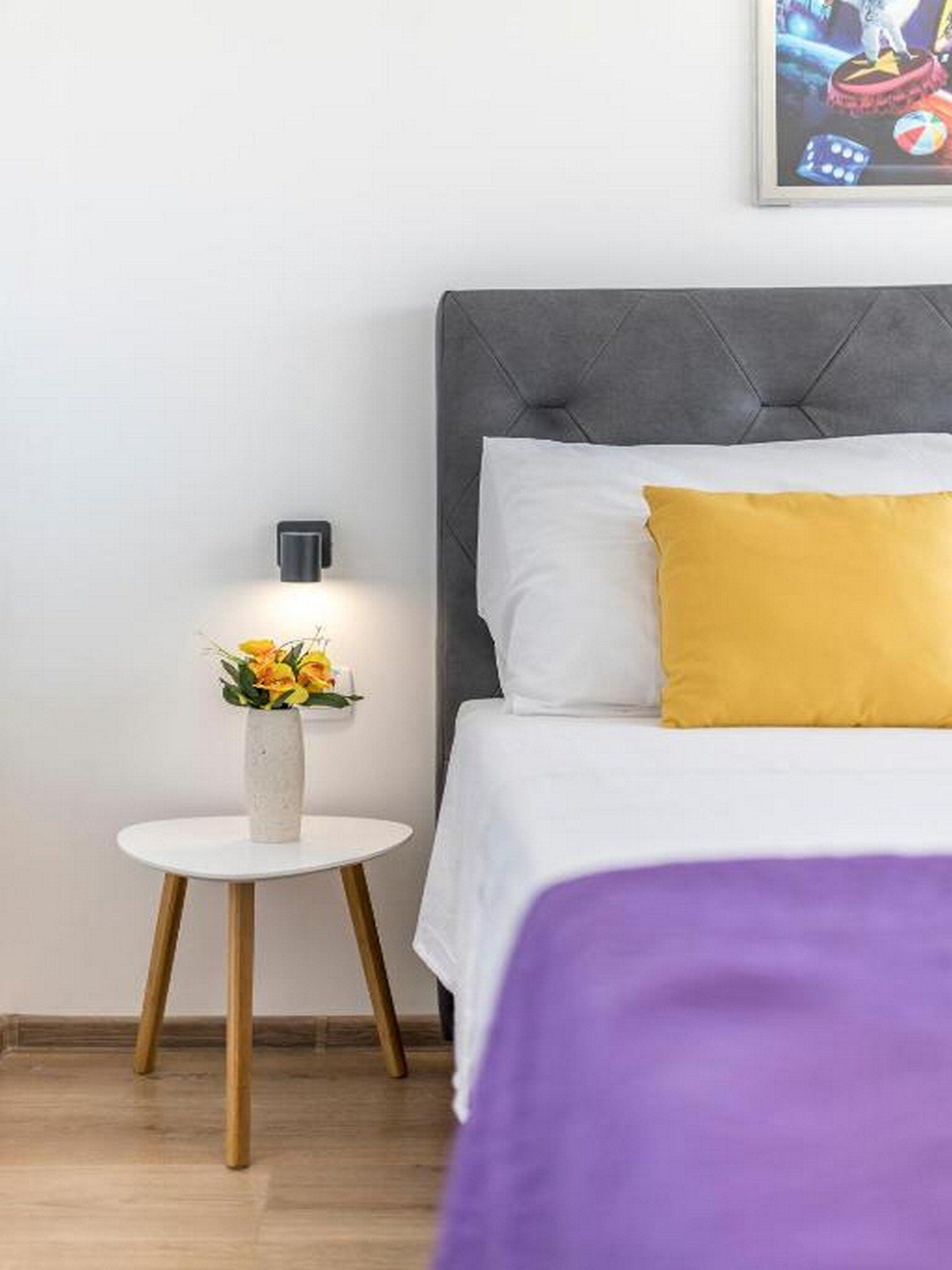 Une chambre d’hôtel bien rangée, avec un lit, une table de chevet ornée d’un vase de fleurs et une lampe fixée au mur.