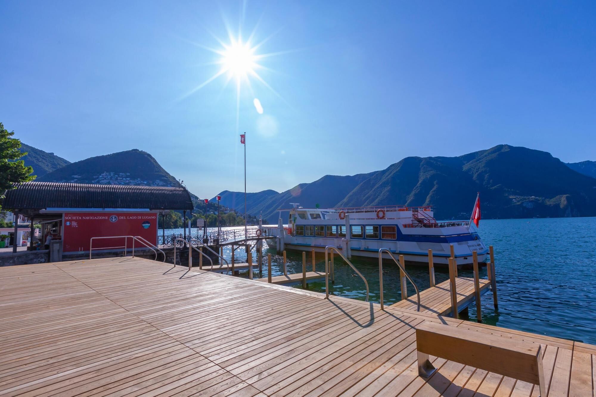A wooden dock with a boat docked at a marina, a red building labeled 'Società Navigazione Del Lago di Lugano', and mountains in the background.