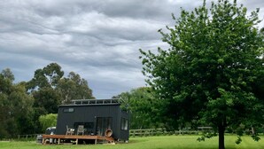 A small, dark-colored cabin with a deck, surrounded by a grassy field and trees. - Merricks North Tiny House (Merricks North)