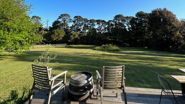 A wooden deck with two chairs and a barrel, overlooking a well-maintained lawn and trees. - Merricks North Tiny House (Merricks North)