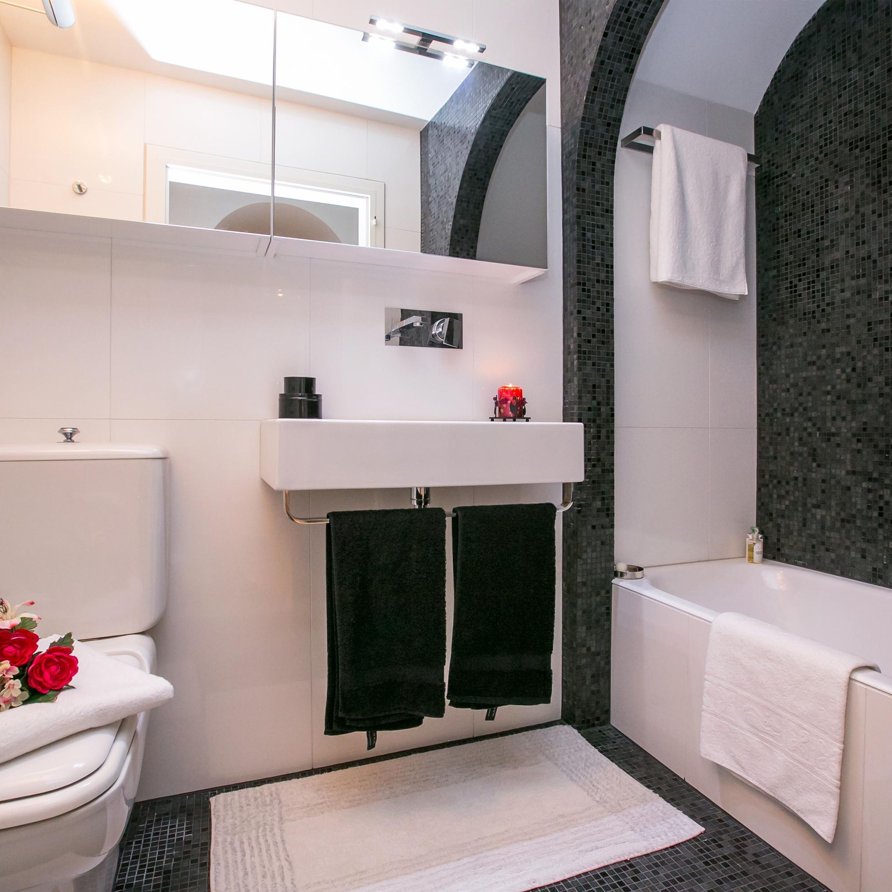 Un baño moderno con inodoro y lavamanos blancos, y una bañera. Hay dos toallas negras colgadas en ganchos, y una pequeña vela roja sobre el lavamanos.