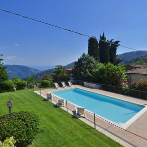 A rectangular swimming pool with a tiled deck, surrounded by a metal fence. There are several white lounge chairs aligned along the poolside. The pool is set against a backdrop of green hills and a clear blue sky. - Villa Medicina in Pescia (Pescia)