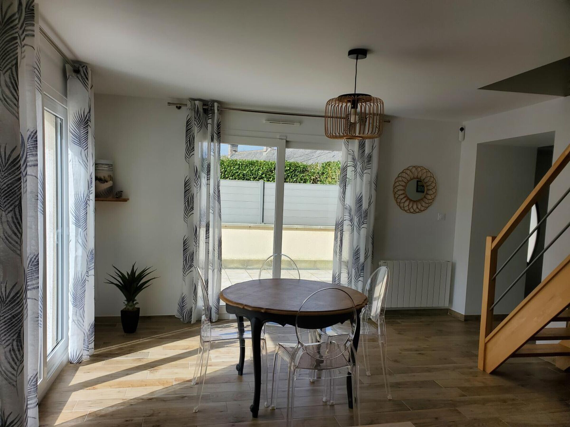 Un comedor con una mesa redonda y sillas, una lámpara colgante, una planta en maceta y vistas a través de un ventanal.
