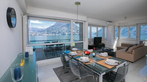 A modern living room with a glass dining table set for four, a sofa, and a TV. Large windows offer a view of a lake and mountains. - Paradiso Incanto in Paradiso Lugano (Paradiso)