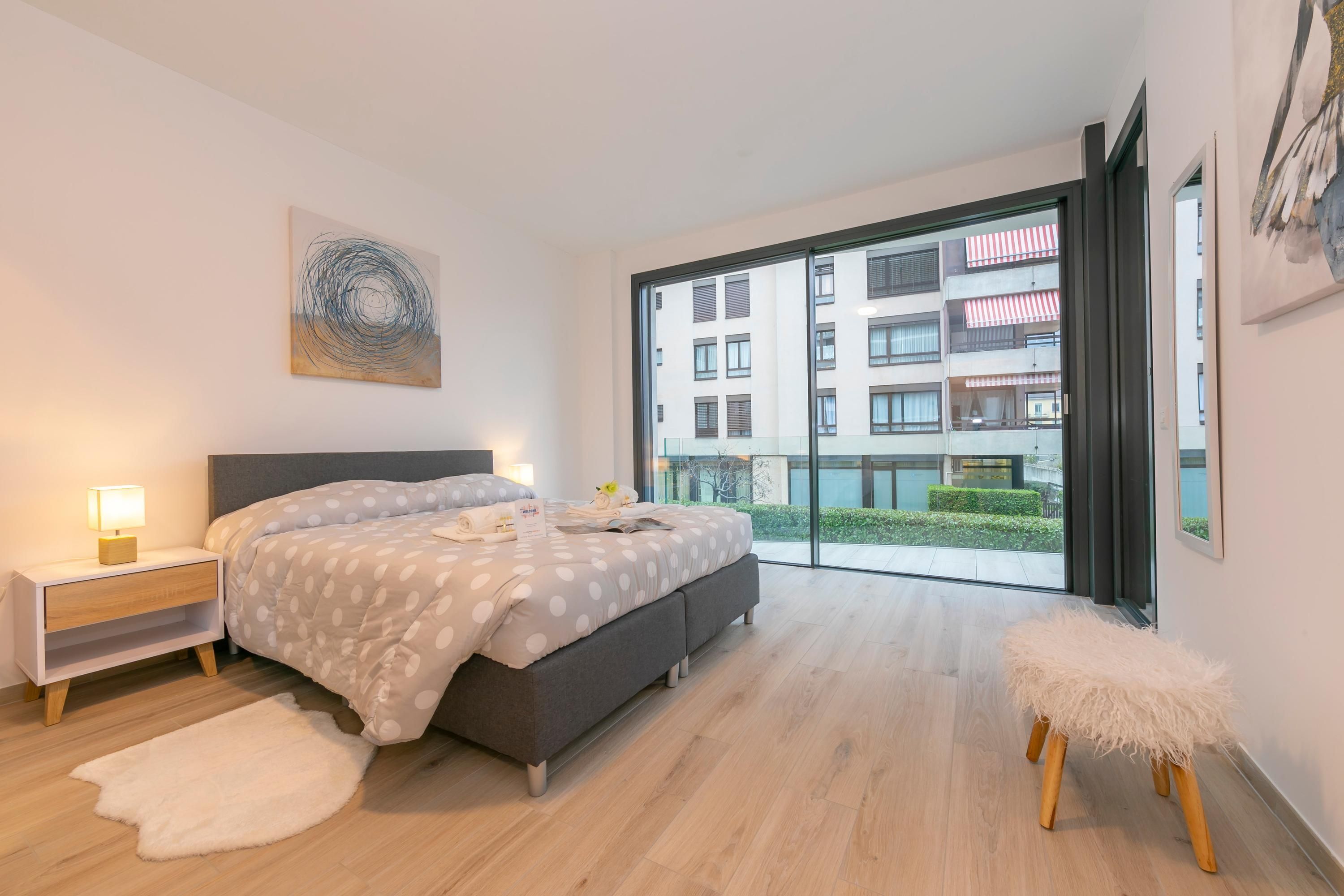 A modern bedroom with a large bed, a nightstand, a fluffy stool, and a view of a building through large windows.