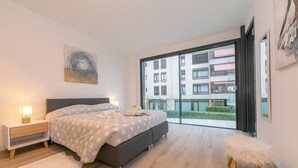 A modern bedroom with a large bed, a nightstand, a fluffy stool, and a view of a building through large windows. - Fairy Home in Viganello Lugano (Lugano)