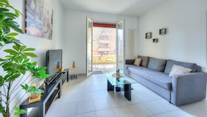 A modern living room with a grey sofa, a glass coffee table, a TV on a black stand, and a large window with a view of a building. - Central Amor in Lugano (Lugano)