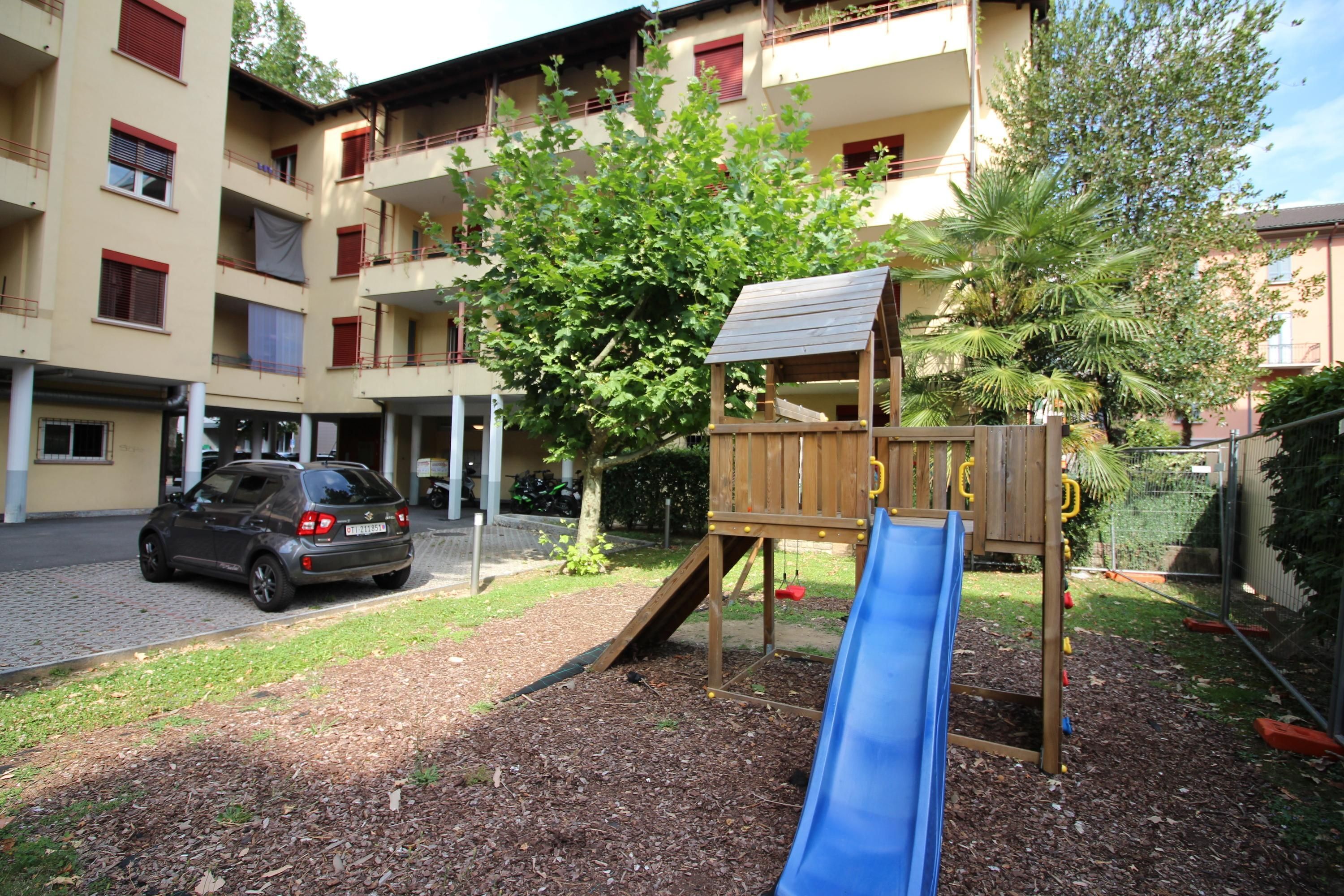 A playground with a slide and climbing structure, surrounded by a courtyard with a parked car and a building with balconies in the background.
