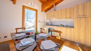 A dining area with a wooden table set for four, a window with a view of a snowy landscape, and wooden kitchen cabinets. - Villa Giorgia Private Pool (Castelveccana)