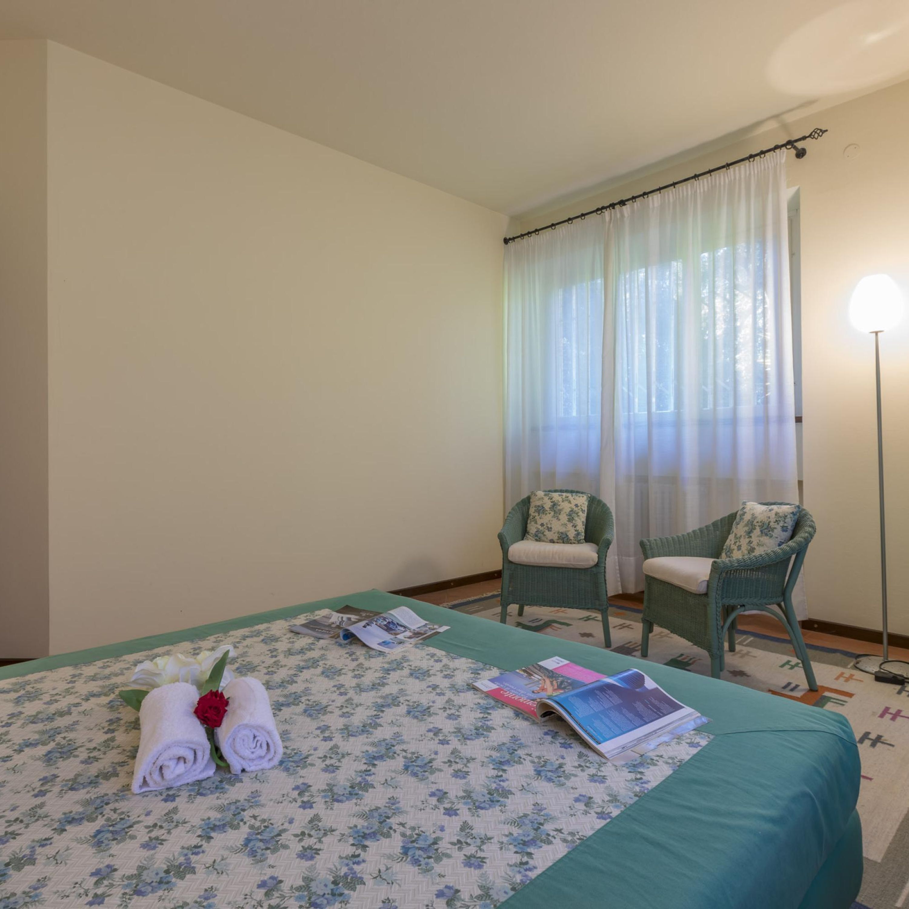 A hotel room with a bed, two chairs, a floor lamp, and a window with sheer curtains.