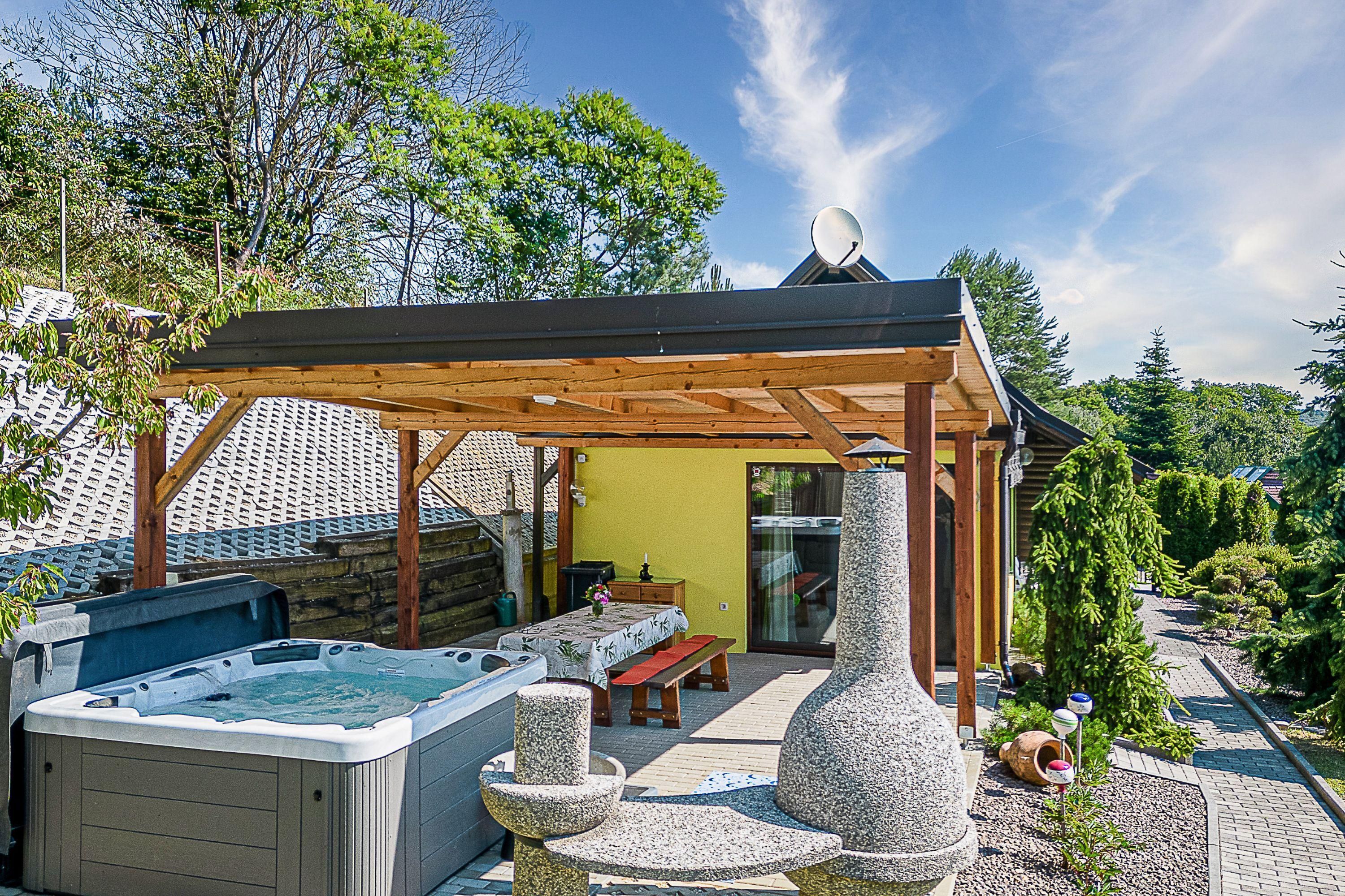 Ein Whirlpool, eine Holzpergola und eine Steinskulptur in einem Garten mit gelber Wand und Holzterrasse.