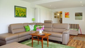 Una sala de estar con un sofá color beige, una mesa de centro de madera y un tapete verde. Hay un cuadro de girasoles en la pared y otro cuadro con flores rojas.