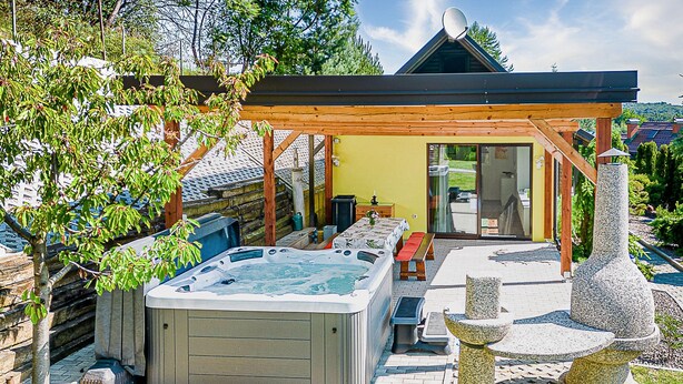 Un jacuzzi bajo una pérgola de madera, una escultura de piedra y una terraza de madera con mobiliario exterior.