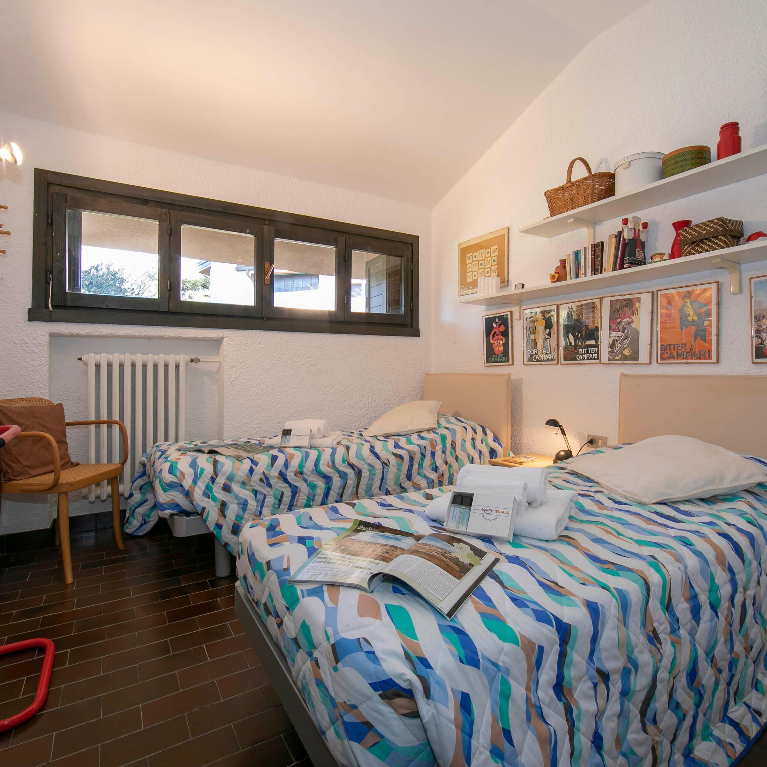 A bedroom with two beds, a window, a radiator, a chair, and shelves with various items.