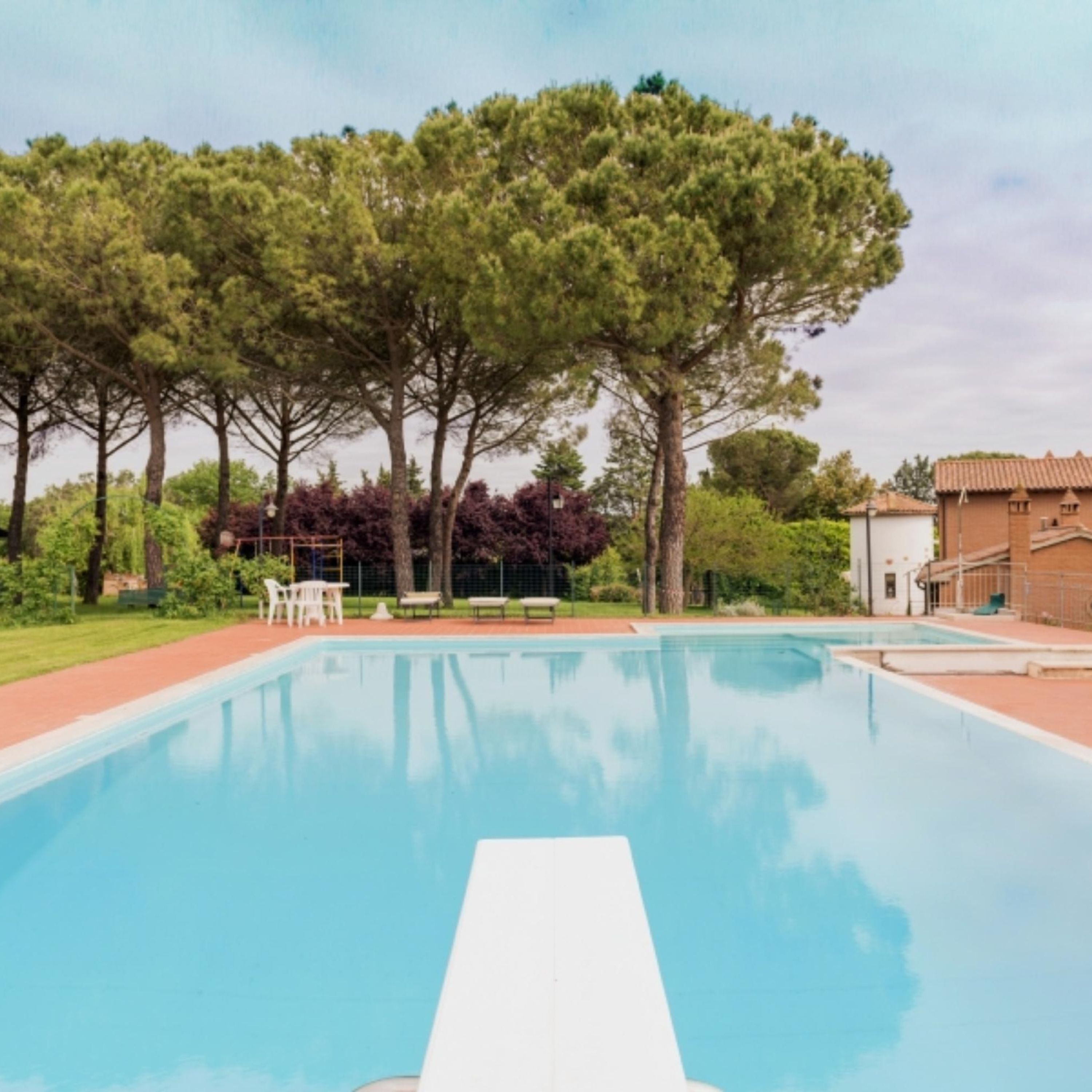 A swimming pool with clear blue water, surrounded by trees and a building with a tiled roof.