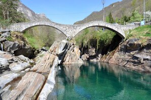 Un pont de pierre enjambe un ruisseau aux eaux claires, bordé de rochers, avec une chaîne de montagnes en arrière-plan.