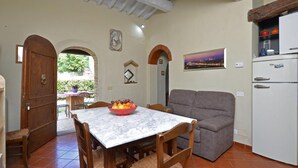 A dining area with a marble table, wooden chairs, and a bowl of fruit. There is a gray sofa and a white refrigerator in the background. - La Badia 2 in Tavarnelle Val di Pesa (Barberino Tavarnelle)