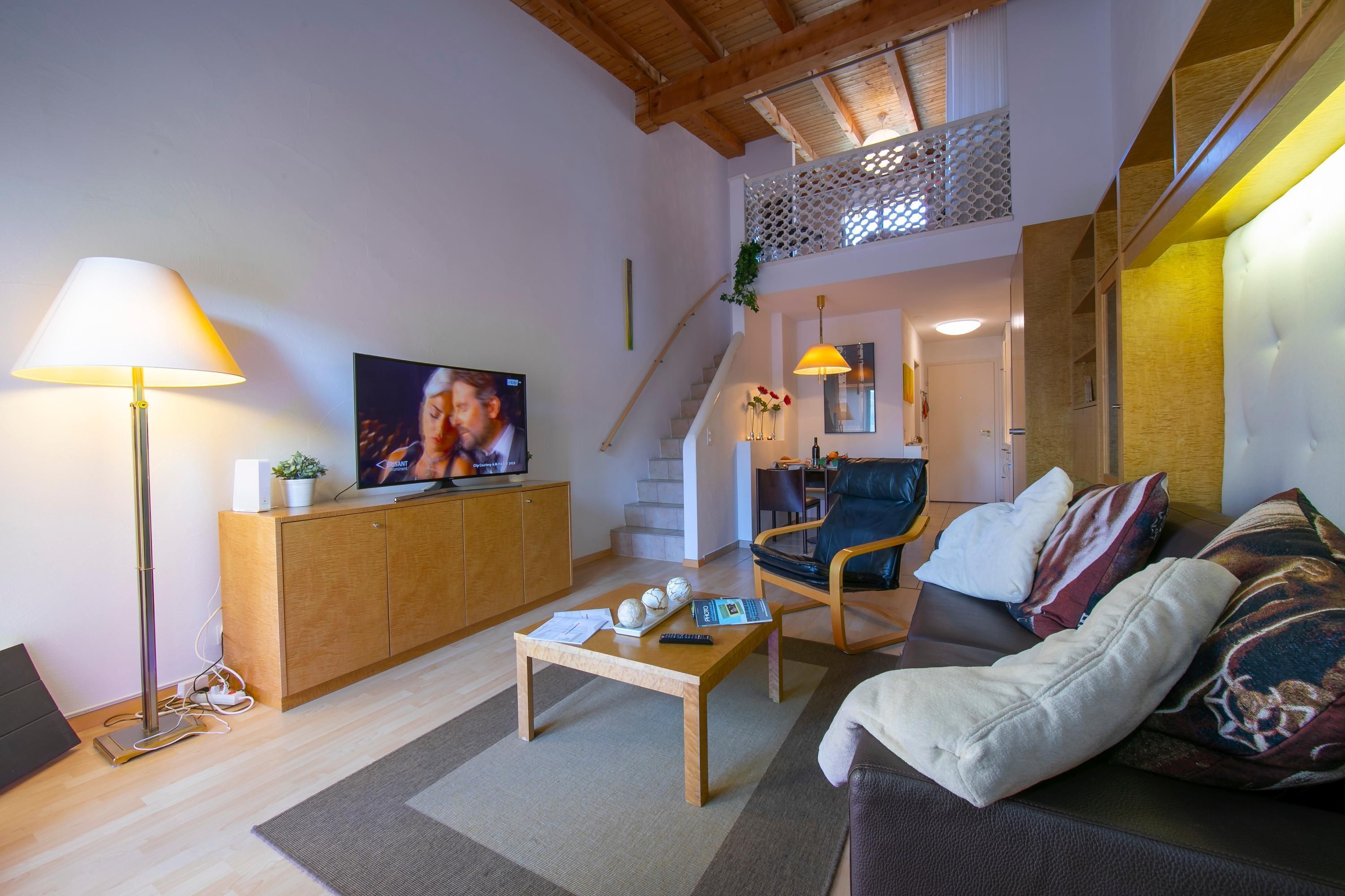 Ein modernes Wohnzimmer mit Sofa, Couchtisch, Fernseher und einer Treppe, die in eine obere Etage führt.