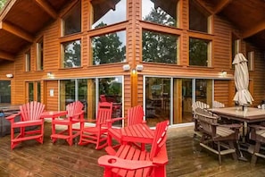A wooden cabin with a large deck, red Adirondack chairs, and a dining table with chairs.