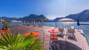 A wooden deck with lounge chairs and umbrellas overlooking a lake with mountains in the background. - Casa Basilico in Lugano (Lugano)