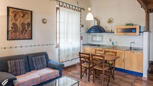 A cozy living room with a sofa, a dining table with chairs, a kitchen area with a stove and cabinets, and a ceiling with exposed beams. - Pomegranate apt With Shared Pool (Castiglione del Lago)