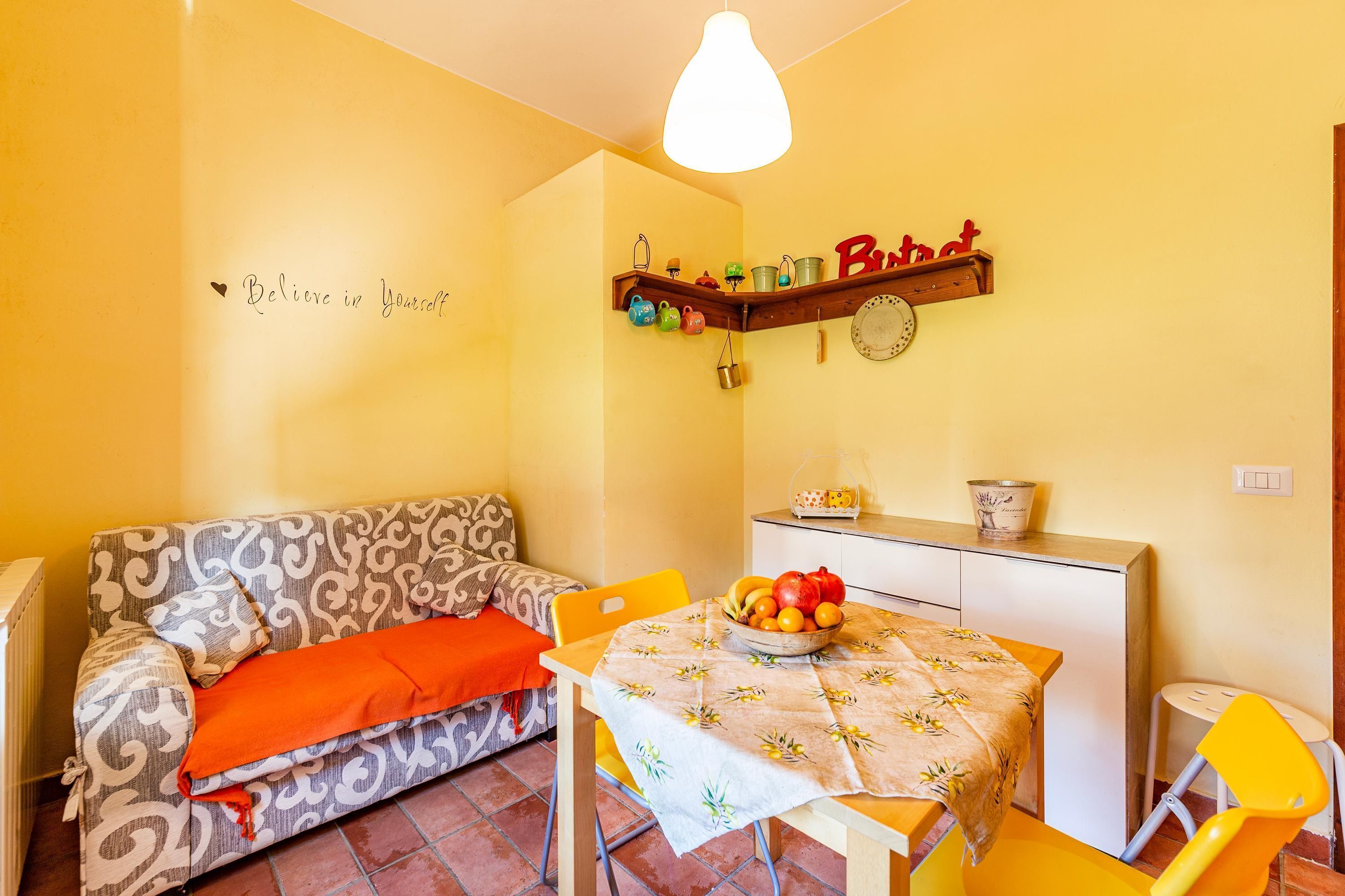 A small, cozy room with a sofa, a dining table with a fruit bowl, and a shelf with kitchen items.
