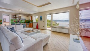 A spacious living room with a large white sofa, a coffee table, and a view of the outdoors through large windows. - Pool Apartment Debeli Rti 3 (Ankaran)