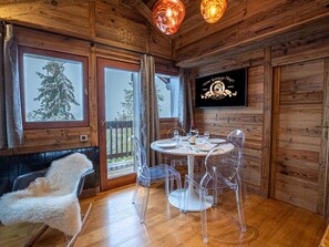 A wooden interior with a round table set for four, clear plastic chairs, a fluffy white chair, and a mounted sign.