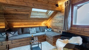Un interior de cabaña acogedor con techo de madera, un sofá con cojines, una mesa de vidrio con una botella y copas, un sillón mullido y un ventanal grande.