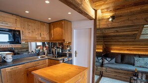 Una cocina acogedora de estilo cabaña con armarios de madera y una isla central, un comedor con sofá y una mesita, y un techo de madera con vigas a la vista.
