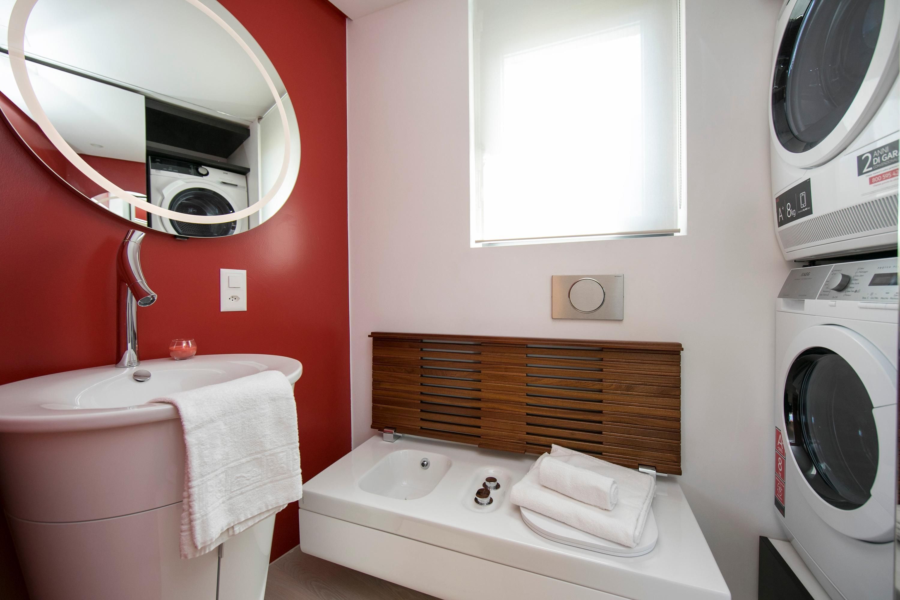 Une salle de bain avec un lavabo rose, un miroir, une tête de lit en bois, une fenêtre et un lave-linge.