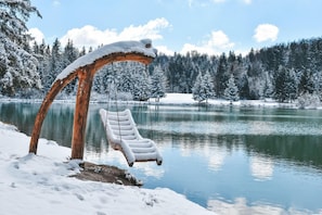 A snow-covered wooden swing chair by a lake with a forest in the background. - Family Friendly Kra Evec Lodge (Bloke)